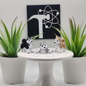 May include: Three small husky figurines in black, white, and brown colors are displayed on a white pedestal. The figurines are arranged on a white cake stand, with two potted plants on either side. A black and white sign is in the background.