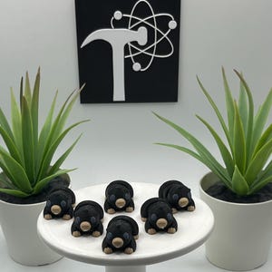 May include: A collection of black bear-shaped confections with tan snouts and paws are arranged on a white cake stand. Two potted green succulent plants flank the stand. A black sign with a white hammer and atom symbol hangs in the background.