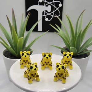 May include: Five small yellow and brown cheetah figurines with black eyes. The figurines are sitting on a white plate.