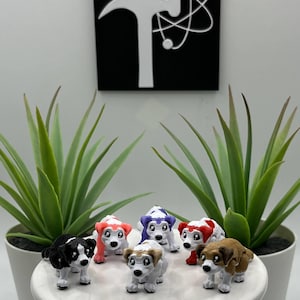May include: A collection of small, colorful dog figurines. Each dog has a white body with accents of black, red, brown, and purple. They are arranged on a white, round pedestal, with green plants in white pots on either side.