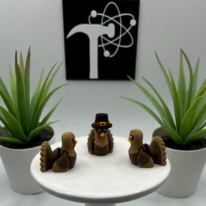 May include: Three small, brown turkey figurines with detailed feathers and beaks are arranged on a white cake stand. One turkey wears a black hat. Two potted green plants flank the stand, and a black and white graphic is in the background.