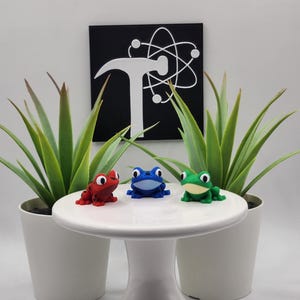 May include: Three colorful frog figurines in red, blue, and green sit on a white, pedestal-style cake stand. Two potted plants flank the stand. A black square with a white hammer and atom symbol hangs in the background.