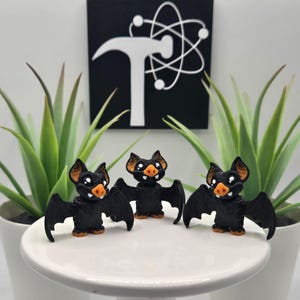 May include: Three black bat figurines with orange noses and feet are displayed on a white pedestal. The bats have outstretched wings and are smiling. The background includes green plants in white pots and a black and white graphic.