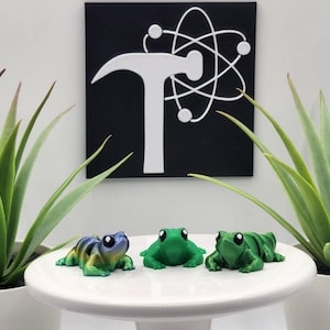 May include: Three small articulated frog figurines in blue-green gradient and solid green colors, featuring large black and white eyes, positioned on a white pedestal. Behind them is a black square sign with a white hammer and atom symbol.