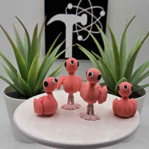 May include: Four pink flamingo figurines with black eyes and light pink feet are arranged on a white cake stand. The background includes green potted plants and a black and white logo.
