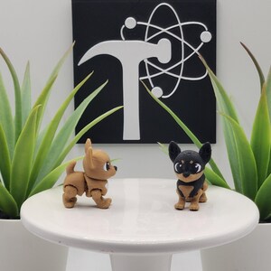 May include: Two small, articulated chihuahua figurines, one tan and one black and tan, are displayed on a white pedestal. The background features a black square with a white hammer and atom symbol, flanked by potted green plants.