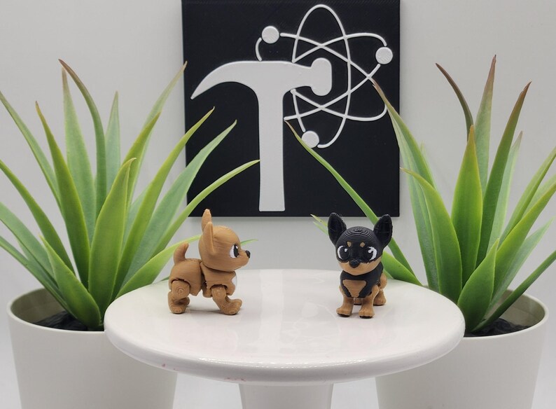 May include: Two small, articulated chihuahua figurines, one tan and one black and tan, are displayed on a white pedestal. The background features a black square with a white hammer and atom symbol, flanked by potted green plants.