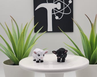 May include: Two small lamb figurines, one white and one black, stand on a white pedestal. Behind them is a black square sign featuring a white hammer icon overlaid with an atom symbol. Two green succulent plants in white pots flank the pedestal.