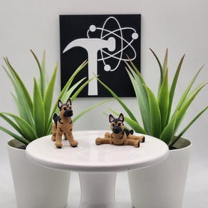 May include: Two small, tan and black German Shepherd figurines on a white pedestal table. One stands, the other reclines. The table is centered between two potted green plants. A black square with a white logo hangs in the background.