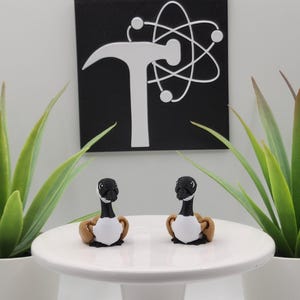 May include: Two small goose figurines with black heads, white chests, and brown wings sit on a white cake stand. The background includes a black square with a white hammer and atom symbol, and two potted green plants.