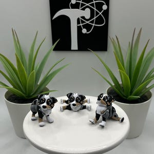 May include: Three miniature dog figurines in black, white, and gray, posed on a white cake stand. Two potted green plants flank the stand. A black square with a white logo is in the background.