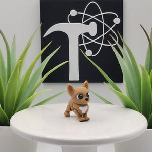 May include: A small, brown Chihuahua figurine with a white chest stands on a white pedestal. The figurine has large, expressive eyes. Two potted green plants flank the pedestal. A black square with a white logo is in the background.