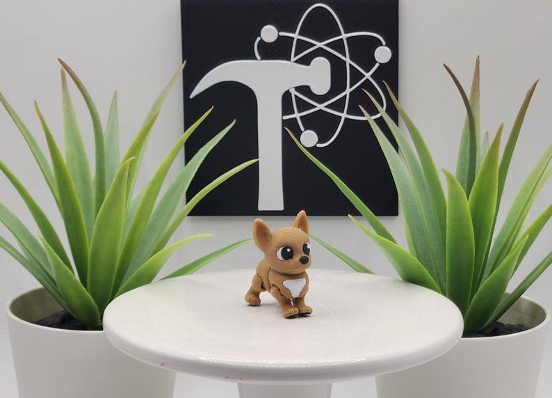 May include: A small, brown Chihuahua figurine with a white chest stands on a white pedestal. The figurine has large, expressive eyes. Two potted green plants flank the pedestal. A black square with a white logo is in the background.