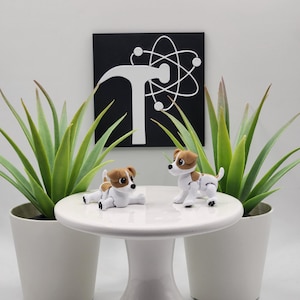 May include: Two Jack Russell Terrier figurines, one lying down and one standing, on a white pedestal table. The background includes two potted plants and a black square with a white hammer and atom symbol.