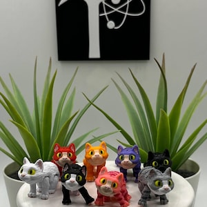 May include: A collection of eight small, multi-colored cat figurines with large yellow eyes, arranged on a white circular stand. The cats feature various patterns and colors including white, red, orange, purple, black, pink, and gray striped designs.