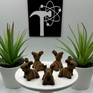 May include: A collection of small, brown moose figurines arranged on a white, round pedestal. The moose have detailed features and are surrounded by potted green plants. A black and white sign is in the background.