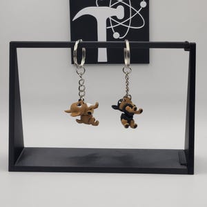 May include: Two dog-shaped keychains hanging from a black display stand. One keychain is brown, the other is black and brown. The keychains have silver-colored hoops and chains. A black square with a white logo is in the background.