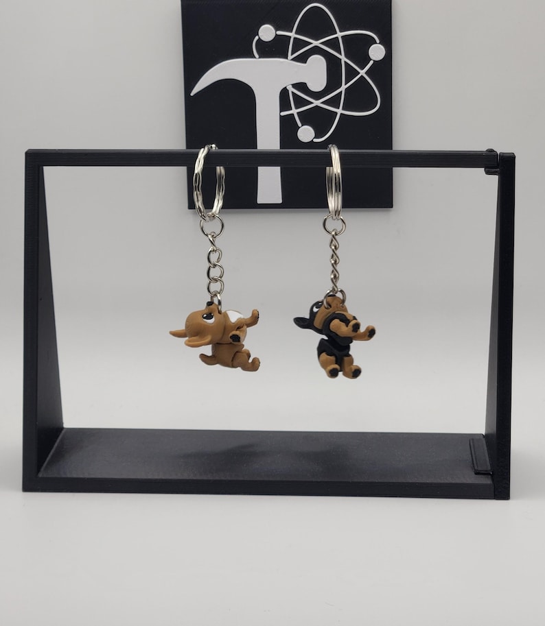 May include: Two dog-shaped keychains hanging from a black display stand. One keychain is brown, the other is black and brown. The keychains have silver-colored hoops and chains. A black square with a white logo is in the background.