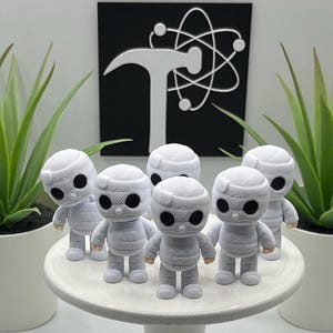 May include: Six white figurines with black eyes and wrapped-style detailing stand on a white cake stand. Two potted green plants flank the figurines. A black square with a white logo is in the background.