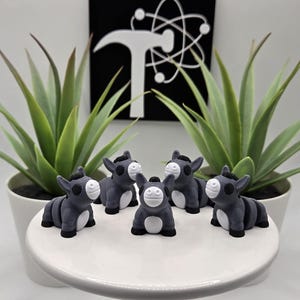 May include: A collection of six gray donkey figurines with white faces and black hooves are arranged on a white, round pedestal. Two potted green plants flank the figurines. A black and white logo is in the background.