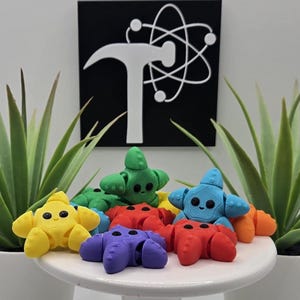 May include: A collection of colorful, star-shaped figurines in yellow, green, red, blue, and purple, each with a smiling face. The figurines are arranged on a white pedestal, with potted plants on either side. A black sign with a white logo is in the background.