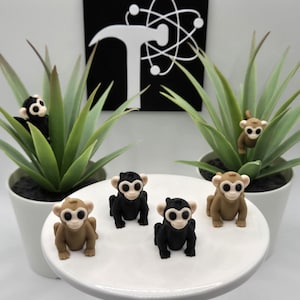 May include: Five small monkey figurines, two brown and three black, with white eyes, sitting on a white cake stand. The monkeys are arranged in a semi-circle.