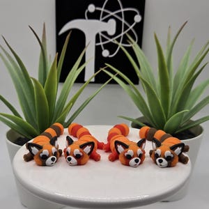 May include: Four orange and brown red panda figurines with white faces and black accents are displayed on a white cake stand. The figurines are lying down. Green plants in white pots are in the background.