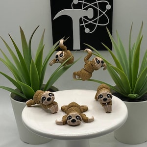 May include: A collection of handmade brown sloth figurines in various poses, some hanging from green plant stems, others resting on a white cake stand. The background includes a black sign with a white hammer and atom symbol, and potted green plants.