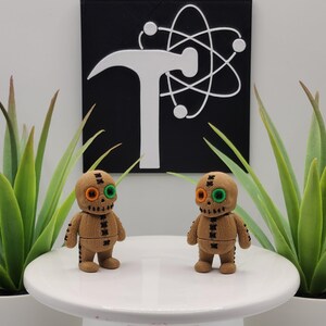 May include: Two tan, textured, 3D-printed figures resembling voodoo dolls with stitched details, one orange and one green button eye each, and a black stitched mouth. They stand on a white surface in front of a black square sign with a white hammer and atom graphic.