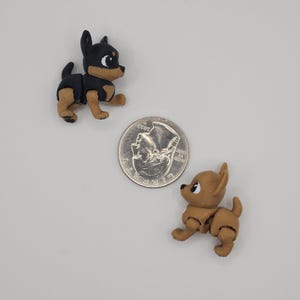 May include: Two small, articulated dog figurines, one black and tan, the other tan, are positioned near a US quarter coin. The black and tan dog has a black body and head with tan legs and chest. The tan dog is a solid tan color.