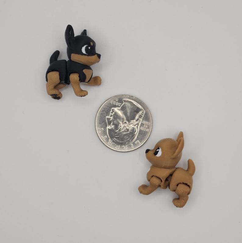 May include: Two small, articulated dog figurines, one black and tan, the other tan, are positioned near a US quarter coin. The black and tan dog has a black body and head with tan legs and chest. The tan dog is a solid tan color.