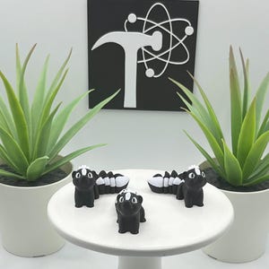 May include: Three small, black skunk figurines with white stripes and accents are arranged on a white cake stand. Two potted green plants flank the stand. A black and white sign with a hammer and atom symbol is in the background.