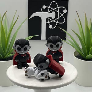 May include: Three small, black and red vampire figurines with gray faces and capes are displayed on a white cake stand. Two green potted plants flank the figurines. A black sign with a white logo is in the background.