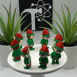May include: A collection of small elf figurines with red hats and green outfits are arranged on a white cake stand. The elves have detailed faces and are surrounded by potted green plants. A black and white graphic is in the background.