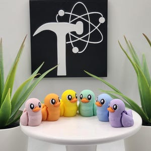 May include: A collection of six colorful bird figurines in pastel shades of pink, orange, yellow, green, blue, and purple. They are arranged on a white table, with a black and white graphic in the background. Two potted plants flank the display.