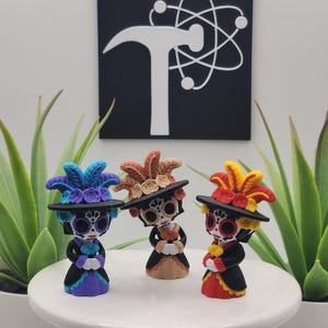 May include: Three small figurines depicting characters with white skull faces, black eye sockets, and floral details. Each wears a wide-brimmed black hat adorned with colorful feathers and flowers, paired with a dress in purple, tan, or red.