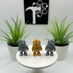 May include: Three small robot figurines in gold and gray, with blue eye details, are displayed on a white cake stand. Two potted green plants flank the figurines. A black and white sign with a hammer and atom symbol is in the background.