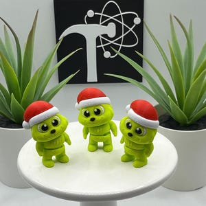 May include: Three lime green figurines wearing red and white Santa hats are displayed on a white, round pedestal. The figurines have large black eyes and are positioned in front of two potted green plants. A black sign with a white logo is in the background.