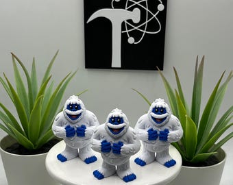 May include: Three white and blue figurines of a smiling snowman-like creature are displayed on a white pedestal. The figurines have blue hands, feet, and facial details. A black and white logo is in the background, and two potted plants flank the display.