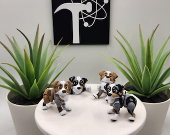 May include: Four small, articulated dog figurines featuring Australian Shepherd-like patterns in brown, white, and grey. The figures are displayed on a white circular stand. A black square sign with a white hammer and atom graphic is visible behind.