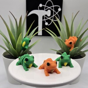 May include: Four small frog figurines in green and orange are displayed on a white surface. Two green frogs and one orange frog are in the foreground, with two more frogs nestled in potted plants in the background. The background features a black and white graphic.