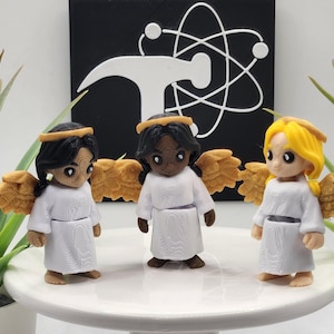 May include: Three small angel figurines with white robes, gold wings, and gold halos. The left figure has black hair, the center figure has black hair and dark skin, and the right figure has yellow hair. Each figure features a textured surface.