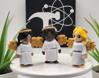 May include: Three small angel figurines with white robes, gold wings, and gold halos. The left figure has black hair, the center figure has black hair and dark skin, and the right figure has yellow hair. Each figure features a textured surface.