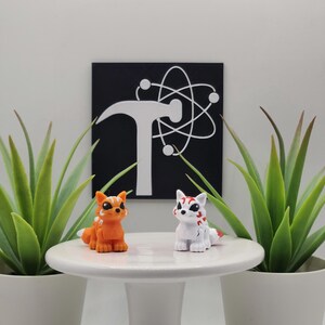 May include: Two small fox figurines, one orange with white stripes and one white with red markings, stand on a white pedestal. Behind them is a black square sign featuring a white hammer icon overlaid with a white orbital atom symbol.