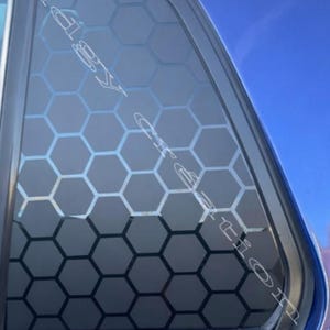 Puede incluir: Una ventana de coche con un patrón de panal de abeja aplicado. El diseño es una serie de hexágonos grises oscuros, creando un efecto geométrico. La ventana está sobre una carrocería azul y un cielo despejado.