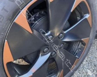 May include: Close-up of a car wheel with a black rim and bronze-colored accents. The tire is black and the wheel has a multi-spoke design. The wheel is partially visible, showing the tire and a portion of the car's body.