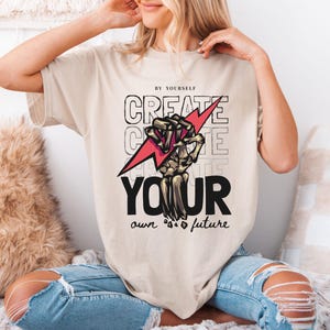 May include: Beige t-shirt with the text "CREATE YOUR own future" in black and white, featuring a skeleton hand holding a red lightning bolt. The shirt is worn by a person wearing ripped blue jeans.