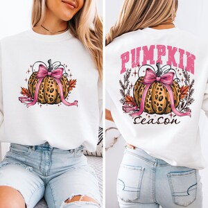 May include: White sweatshirt with a pumpkin design and a pink bow. The pumpkin has a leopard print pattern, and the text "Pumpkin Season" is displayed. The design is repeated on the front and back of the sweatshirt.