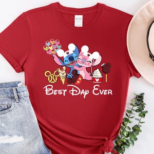 A red t-shirt with a Disney-themed graphic and text, along with a pair of jeans and a hat, all laid out on a white surface.