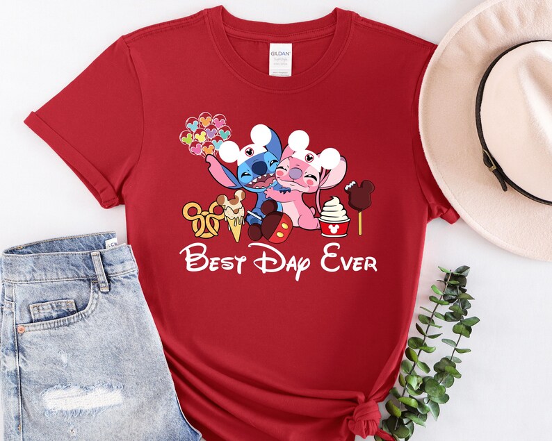 A red t-shirt with a Disney-themed graphic and text, along with a pair of jeans and a hat, all laid out on a white surface.